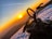 A snowmobile is parked on a snowy terrain during a vibrant sunset. The sky is an array of warm hues with orange and yellow, while the snow reflects some of the sunlight. There are contrails in the sky, suggesting passing airplanes, and the scene conveys a serene and adventurous atmosphere.