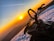 A snowmobile is parked on a snowy terrain during a vibrant sunset. The sky is an array of warm hues with orange and yellow, while the snow reflects some of the sunlight. There are contrails in the sky, suggesting passing airplanes, and the scene conveys a serene and adventurous atmosphere.