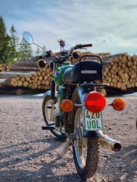 A vintage motorcycle with the brand name 'Simson' is parked on a gravel surface. The motorcycle has a green and black color scheme with a visible license plate reading '420 UOL'. In the background, there are stacks of cut logs, and a person can be seen standing near them.