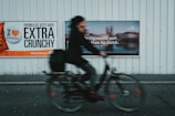 A blurred cyclist rides past two large advertisements on a metal wall. The left advertisement features a bold message about paprika chips being extra crunchy, with a color scheme of orange, black, and white. The right advertisement promotes a high-speed internet offer for Zurich, displaying a mix of blurred imagery and text.