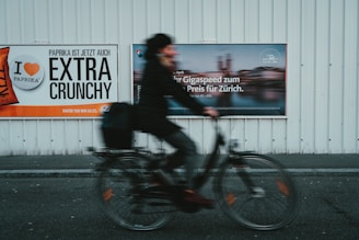 A blurred cyclist rides past two large advertisements on a metal wall. The left advertisement features a bold message about paprika chips being extra crunchy, with a color scheme of orange, black, and white. The right advertisement promotes a high-speed internet offer for Zurich, displaying a mix of blurred imagery and text.
