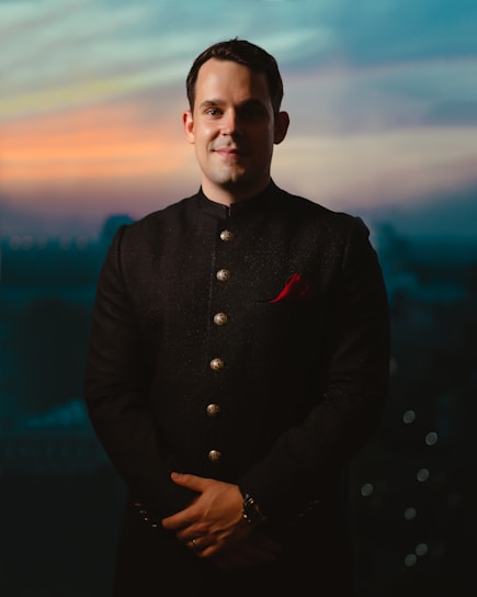 A close-up of a man wearing a sleek black thobe with subtle embroidery, standing confidently against a city backdrop at dusk.