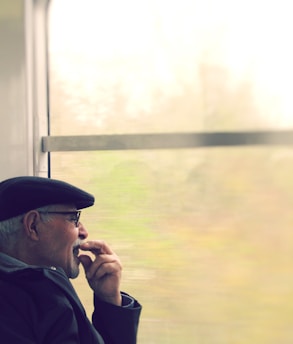An older man wearing a flat cap and glasses is sitting thoughtfully by a window. He is looking outside with his hand resting near his mouth, appearing deep in contemplation. The view outside the window is blurred, suggesting motion, and the background is light and airy, possibly indicating daylight.