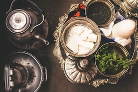A decorative silver tray holds an assortment of items, including a bowl of white cheese, fresh mint leaves, loose tea, and a white item wrapped in paper. A metallic tea kettle sits next to the tray.