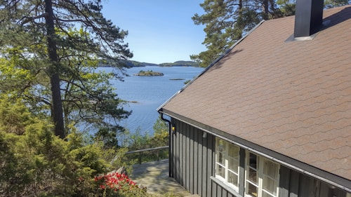 A serene lakeside view featuring a grey wooden house with a sloped roof. Pine trees frame the scene, and a vibrant patch of red flowers contrasts with the greenery. The calm water of the lake reflects the clear blue sky, while small islands can be seen in the distance.