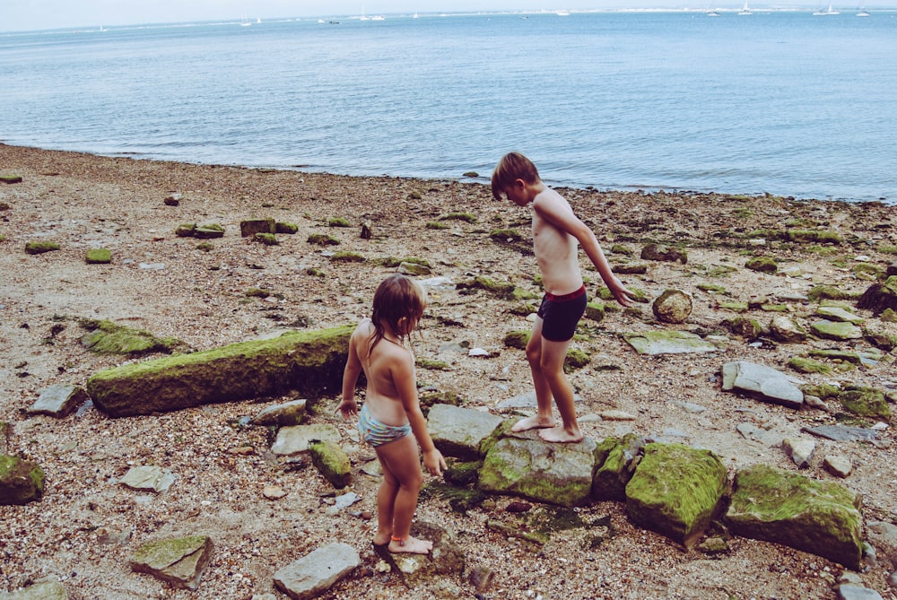 two children walking on sand with rocks near body of water