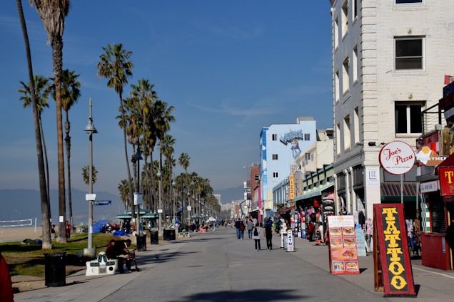 A lively beachfront promenade is lined with tall palm trees on one side and colorful buildings housing various shops and eateries on the other. People are strolling along the walkway, some stopping to look at signs advertising food and tattoos. The sky is clear and blue, with a hint of mountains in the distance.