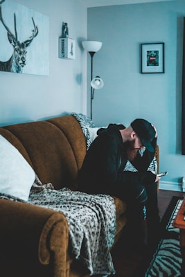 A person in dark clothing and a cap is sitting on a brown sofa, looking at a smartphone. The room has a cozy feel with a patterned throw on the sofa and a floor lamp nearby. A piece of wall art featuring a stag is visible, along with a framed picture on the wall.