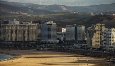 A coastal cityscape with a row of high-rise buildings in front of rolling hills. In the foreground, a sandy beach with a few people walking or standing. The background features wind turbines on the distant hills under a cloudy sky.