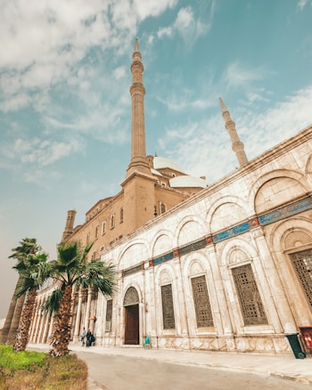 A grand mosque with tall minarets and an ornate fa&ccedil;ade, featuring intricate architectural details and a series of arched windows. Palm trees line the pathway, adding a touch of greenery against the backdrop of a blue sky with scattered clouds.