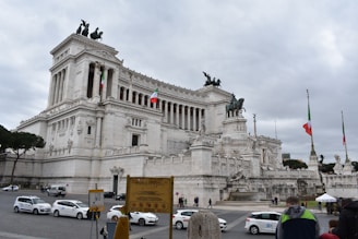 A grand, ornate marble building featuring classical architectural elements such as columns and statues, situated in a public square. Italian flags are visible, and several people and cars are present around the area. The sky is overcast with a hint of grey.