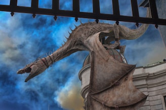 A large, menacing dragon with scales and sharp teeth perched on the roof of a building, with its wings partially spread. The sky behind is filled with dramatic clouds in shades of blue and gray, enhancing the mystical atmosphere.