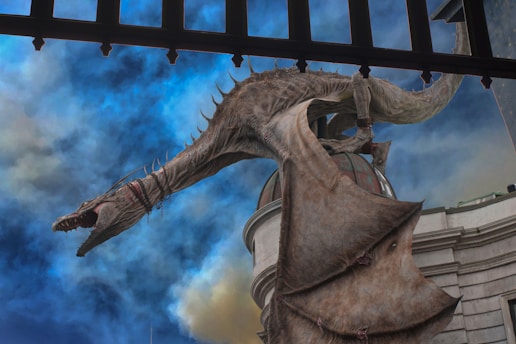 A large, menacing dragon with scales and sharp teeth perched on the roof of a building, with its wings partially spread. The sky behind is filled with dramatic clouds in shades of blue and gray, enhancing the mystical atmosphere.