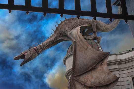 A large, menacing dragon with scales and sharp teeth perched on the roof of a building, with its wings partially spread. The sky behind is filled with dramatic clouds in shades of blue and gray, enhancing the mystical atmosphere.