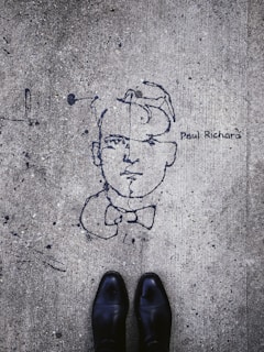 Graffiti art on a concrete surface features a minimalistic line drawing of a face with a bow tie, labeled with the name 'Paul Richard'. The image includes a pair of black shoes at the bottom, indicating a person standing above the artwork.