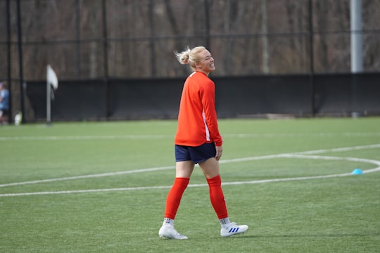 A person in sports attire is standing on a soccer field, wearing a red long-sleeve shirt, navy shorts, red socks, and white athletic shoes. The individual appears to be smiling and is positioned near the center of a green pitch with cones marking areas on the field. The background shows a fenced area with trees.