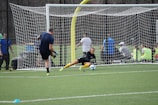 A soccer goalkeeper dives to block a shot during practice on a green turf field. The goalkeeper is wearing yellow gloves and matching knee-high socks, while the player taking the shot is wearing a blue shirt and black shorts. In the background, a group of people are watching from behind a netted fence.