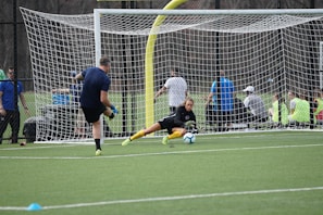 A soccer goalkeeper dives to block a shot during practice on a green turf field. The goalkeeper is wearing yellow gloves and matching knee-high socks, while the player taking the shot is wearing a blue shirt and black shorts. In the background, a group of people are watching from behind a netted fence.