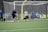 A soccer goalkeeper dives to block a shot during practice on a green turf field. The goalkeeper is wearing yellow gloves and matching knee-high socks, while the player taking the shot is wearing a blue shirt and black shorts. In the background, a group of people are watching from behind a netted fence.