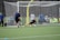 A soccer goalkeeper dives to block a shot during practice on a green turf field. The goalkeeper is wearing yellow gloves and matching knee-high socks, while the player taking the shot is wearing a blue shirt and black shorts. In the background, a group of people are watching from behind a netted fence.