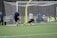 A soccer goalkeeper dives to block a shot during practice on a green turf field. The goalkeeper is wearing yellow gloves and matching knee-high socks, while the player taking the shot is wearing a blue shirt and black shorts. In the background, a group of people are watching from behind a netted fence.
