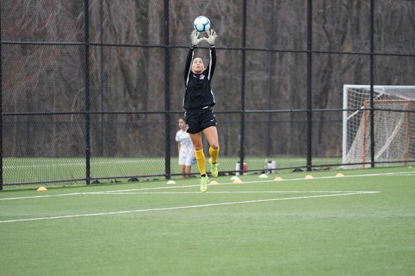A person wearing a black sports outfit and yellow socks is leaping into the air with arms raised to catch a soccer ball on a green field. The soccer field is enclosed by a tall black fence, and an empty goalpost is visible in the background. A few cones are lined up on the ground near the fence, and another person stands nearby.