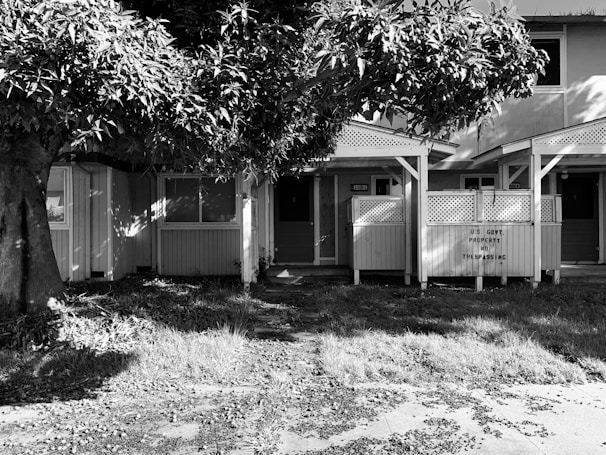 A black and white photo shows a small building with two front doors, partially obscured by a large tree. The building has two porches with lattice work, and the sign on one porch reads 'U.S. Govt. Property No Trespassing'. The ground is covered with grass and leaves, suggesting an untended or abandoned property.