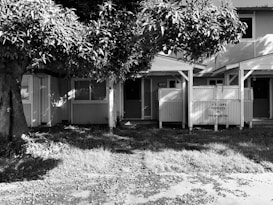 A black and white photo shows a small building with two front doors, partially obscured by a large tree. The building has two porches with lattice work, and the sign on one porch reads 'U.S. Govt. Property No Trespassing'. The ground is covered with grass and leaves, suggesting an untended or abandoned property.