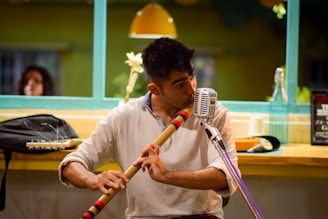 A musician is playing a wooden flute while seated at a table. A vintage microphone is positioned in front of him. On the table, there are various items such as a plastic bottle, a cup, and a small black bag, against a backdrop of a brightly lit cafe with green window frames and a hanging lamp.