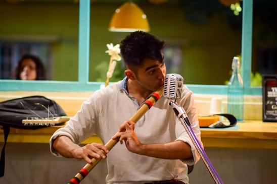 A musician is playing a wooden flute while seated at a table. A vintage microphone is positioned in front of him. On the table, there are various items such as a plastic bottle, a cup, and a small black bag, against a backdrop of a brightly lit cafe with green window frames and a hanging lamp.