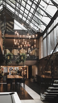 A modern interior space with large glass windows and a geometric ceiling structure. Hanging light bulbs create a sophisticated ambiance. A long wooden table is surrounded by dark chairs, and plants are suspended above. A staircase with metallic railings leads to an upper level.