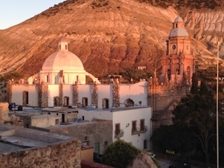 A historic town features an impressive architectural scene with a large dome and a clock tower bathed in warm sunset light. The buildings have a classic stone and stucco construction, and the background displays a rugged, mountainous terrain.