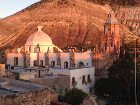 A historic town features an impressive architectural scene with a large dome and a clock tower bathed in warm sunset light. The buildings have a classic stone and stucco construction, and the background displays a rugged, mountainous terrain.