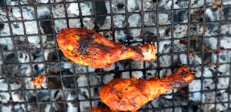 Grilled chicken drumsticks with a rich, charred texture resting on a metal barbecue grill. Ashes and charcoal can be seen underneath the grill grates.