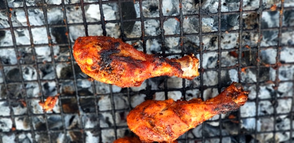 Grilled chicken drumsticks with a rich, charred texture resting on a metal barbecue grill. Ashes and charcoal can be seen underneath the grill grates.