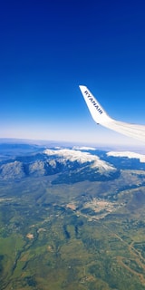 An aerial view features a plane's wing with a city below, surrounded by expansive green fields and snowy mountains in the distance, under a clear blue sky.