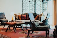 A cozy living room features a mix of traditional and modern furniture. There's a patterned carpet, a decorative screen with intricate patterns, and cushioned chairs placed around a coffee table. Lamps with warm lighting are situated on either side of a sofa adorned with various throw pillows.