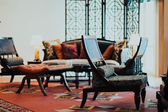 A cozy living room features a mix of traditional and modern furniture. There's a patterned carpet, a decorative screen with intricate patterns, and cushioned chairs placed around a coffee table. Lamps with warm lighting are situated on either side of a sofa adorned with various throw pillows.