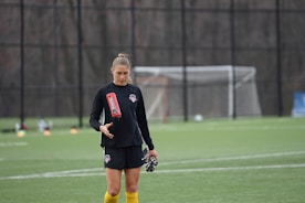 A person standing on a soccer field holding a water bottle, wearing a black sports uniform with yellow socks. In the background, there is a soccer goal and some equipment on the grass.