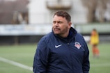 A man wearing a navy blue sports jacket is standing outdoors on a sports field. The jacket has a logo on it, and there is a blurred figure in sports attire in the background.