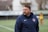 A man wearing a navy blue sports jacket is standing outdoors on a sports field. The jacket has a logo on it, and there is a blurred figure in sports attire in the background.