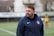 A man wearing a navy blue sports jacket is standing outdoors on a sports field. The jacket has a logo on it, and there is a blurred figure in sports attire in the background.