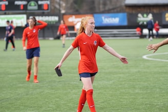 A soccer player in a red jersey with navy shorts is extending her arms, possibly for a handshake or high-five. She stands on a soccer field with a blurred background showing a scoreboard and other players in similar red jerseys. One player in focus has her hand on her head as she walks.