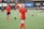 A soccer player in a red jersey with navy shorts is extending her arms, possibly for a handshake or high-five. She stands on a soccer field with a blurred background showing a scoreboard and other players in similar red jerseys. One player in focus has her hand on her head as she walks.