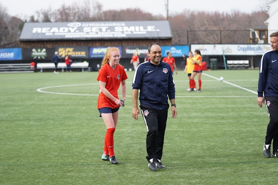 A group of people are on a soccer field, with some wearing sports attire. In the foreground, a person in red sports clothing is walking alongside someone in a dark tracksuit. Other people are visible in the background, some wearing yellow jerseys. A building with advertising banners, including a prominent sign reading 'Ready, Set, Smile,' is in the background.