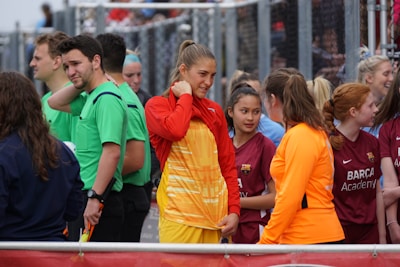A group of young people, some dressed in sports uniforms, are gathered together. The scene appears to be at a sports event, possibly a soccer match, with several individuals wearing shirts labeled 'Barça Academy.' One person in a bright orange top stands out, engaging in conversation with another in a bright red and yellow outfit. Referees in green uniforms are also present in the background.