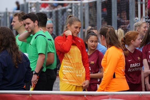 A group of young people, some dressed in sports uniforms, are gathered together. The scene appears to be at a sports event, possibly a soccer match, with several individuals wearing shirts labeled 'Barça Academy.' One person in a bright orange top stands out, engaging in conversation with another in a bright red and yellow outfit. Referees in green uniforms are also present in the background.