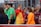 A group of young people, some dressed in sports uniforms, are gathered together. The scene appears to be at a sports event, possibly a soccer match, with several individuals wearing shirts labeled 'Barça Academy.' One person in a bright orange top stands out, engaging in conversation with another in a bright red and yellow outfit. Referees in green uniforms are also present in the background.
