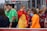 A group of young people, some dressed in sports uniforms, are gathered together. The scene appears to be at a sports event, possibly a soccer match, with several individuals wearing shirts labeled 'Barça Academy.' One person in a bright orange top stands out, engaging in conversation with another in a bright red and yellow outfit. Referees in green uniforms are also present in the background.