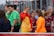 A group of young people, some dressed in sports uniforms, are gathered together. The scene appears to be at a sports event, possibly a soccer match, with several individuals wearing shirts labeled 'Barça Academy.' One person in a bright orange top stands out, engaging in conversation with another in a bright red and yellow outfit. Referees in green uniforms are also present in the background.